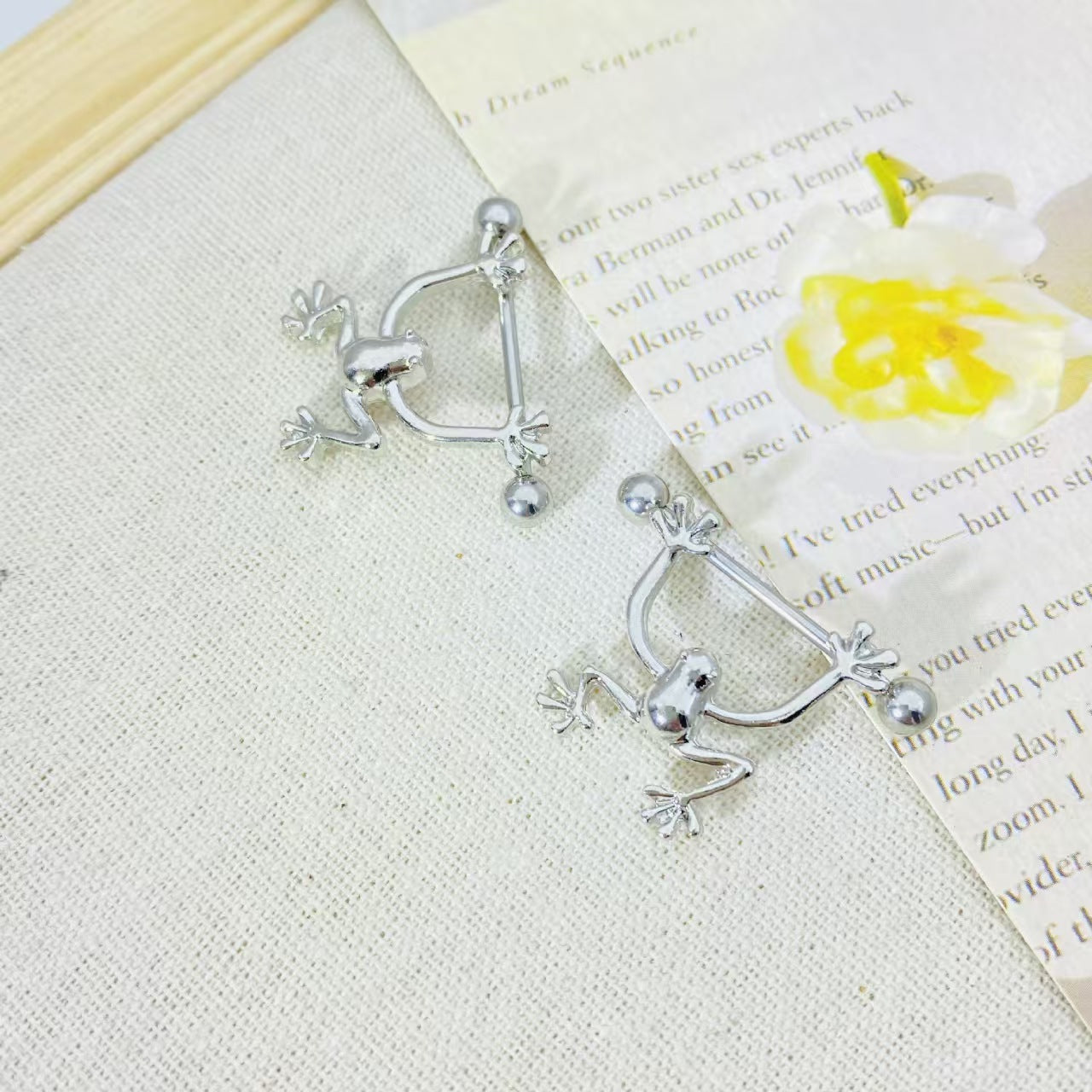 Flat lay of frog nipple jewelry resting on a book with a yellow flower for aesthetic presentation.
