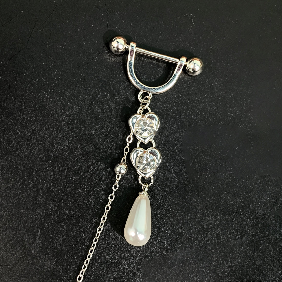 Close-up detailed view of the barbell piercing showing double heart-shaped accents with glittering stones and a smooth teardrop dangle pendant.