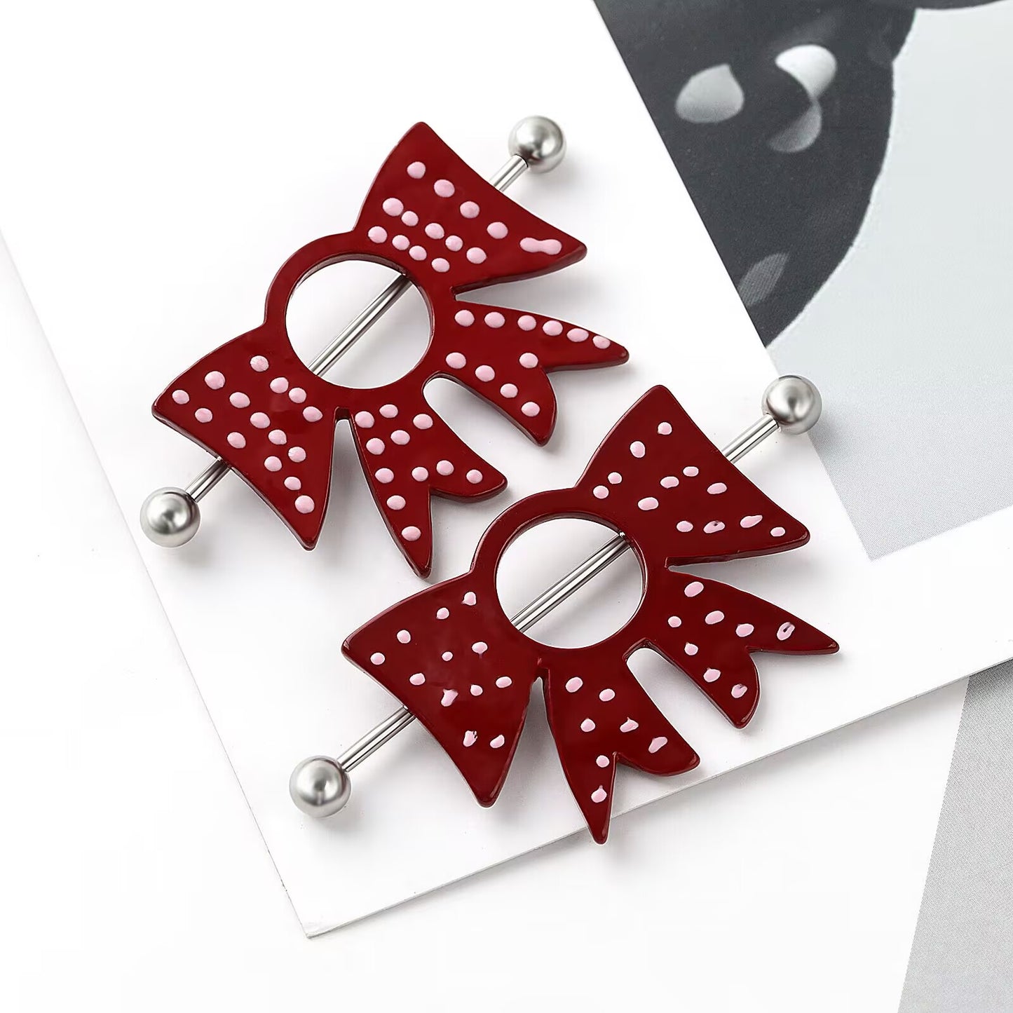 Close up flatlay view of trendy coquette aesthetic red bow nipple shields with 14G barbells for alternative body piercing.