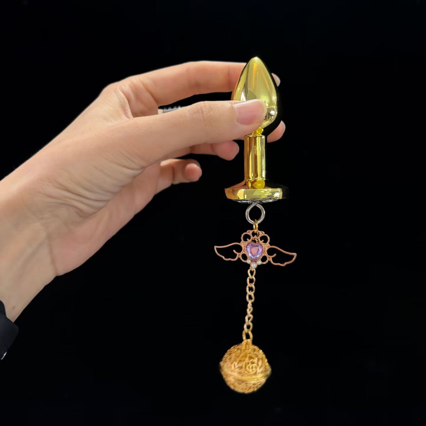 Hand holding a polished gold metal anal plug with dangling purple heart winged pendant and filigree sphere.