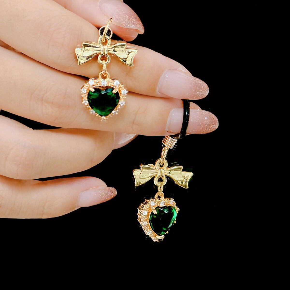 Close-up of a hand holding a pair of non-pierced nipple rings with green heart gemstones and gold bows.