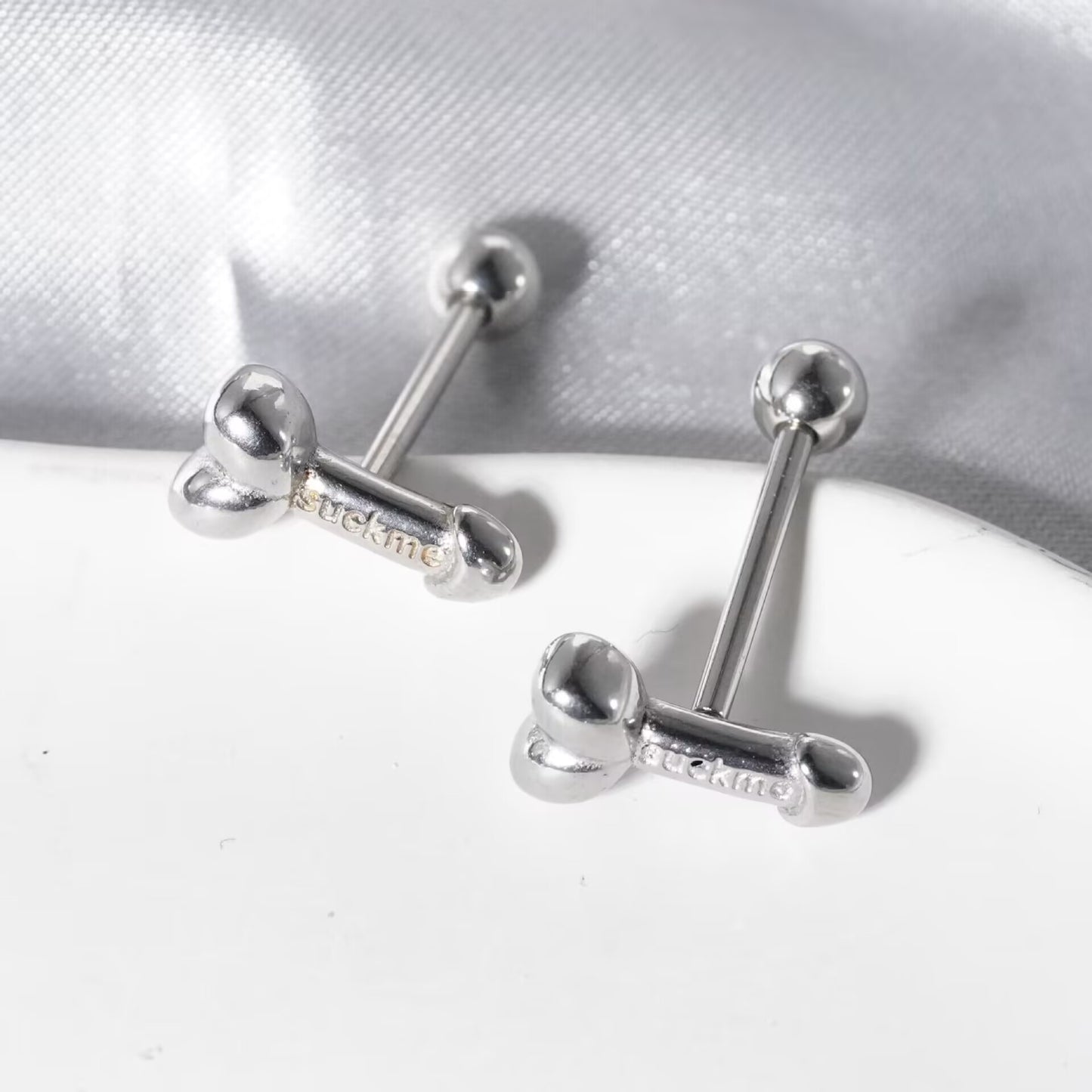 Close-up of silver penis-shaped tongue rings with engraved suck me lettering resting on a display tray.