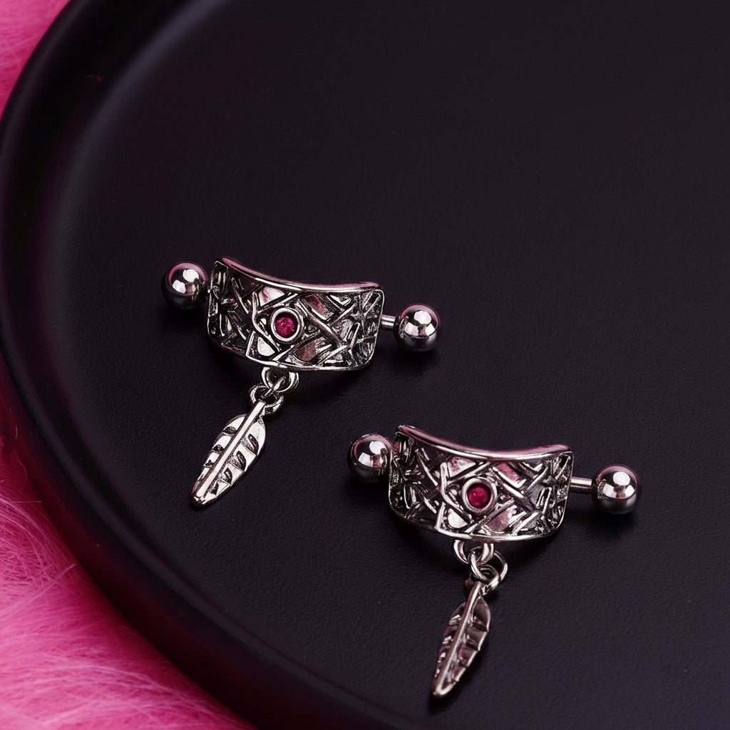 A pair of decorative boho nipple shields with red accents and leaf pendants displayed on a black matte tray with a pink plush background, showing the jewelry's curved fit.