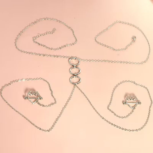 Flat lay product photo of a silver triple O-ring choker body chain connected to a pair of rhinestone-embellished hollow heart nipple shields over a soft pink background.