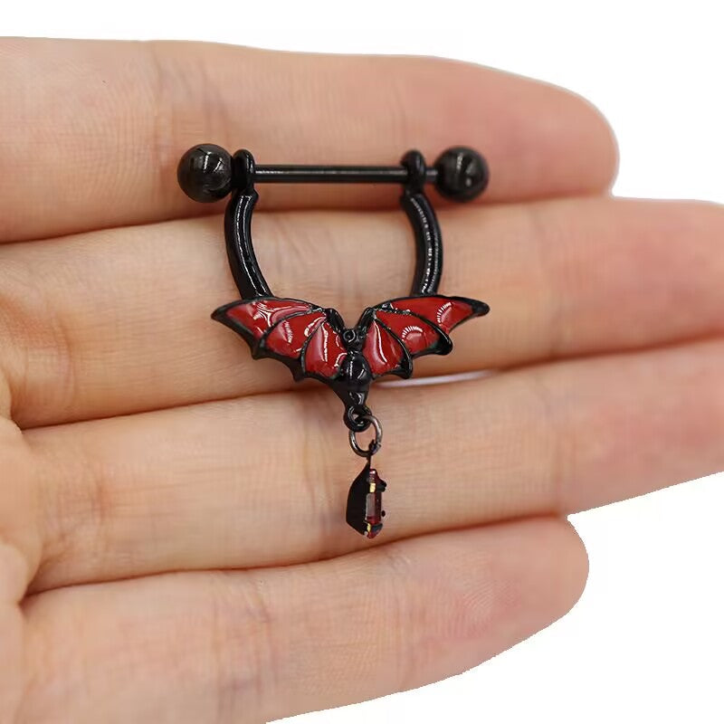 A close-up shot of a hand holding a gothic black and red bat nipple shield piercing with a blood drop charm to show real-life detail.