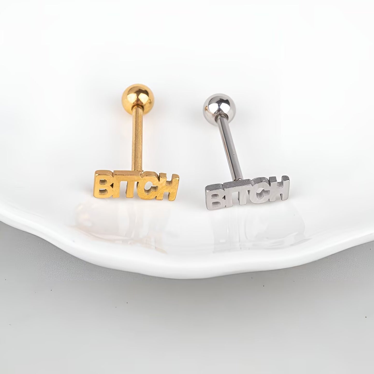 Pair of gold and silver BITCH letter tongue studs on white plate background.