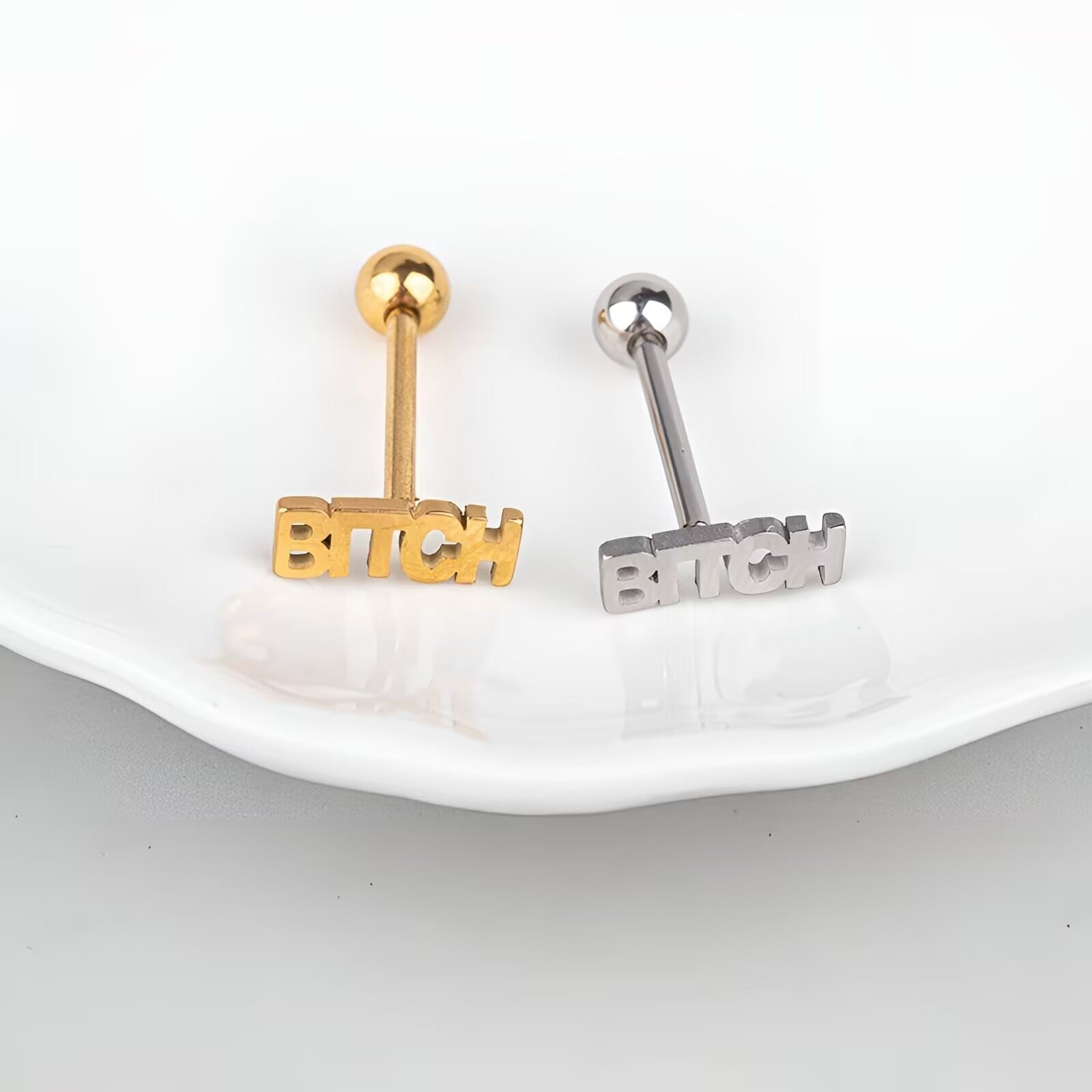 Pair of gold and silver BITCH letter tongue studs on white plate background.