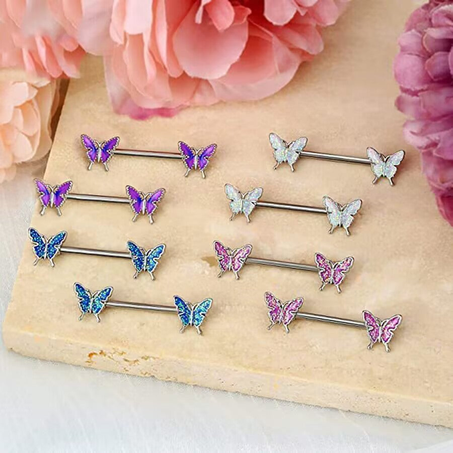 Four pairs of colorful glitter butterfly nipple barbells arranged on a beige stone slab surrounded by pink flowers.