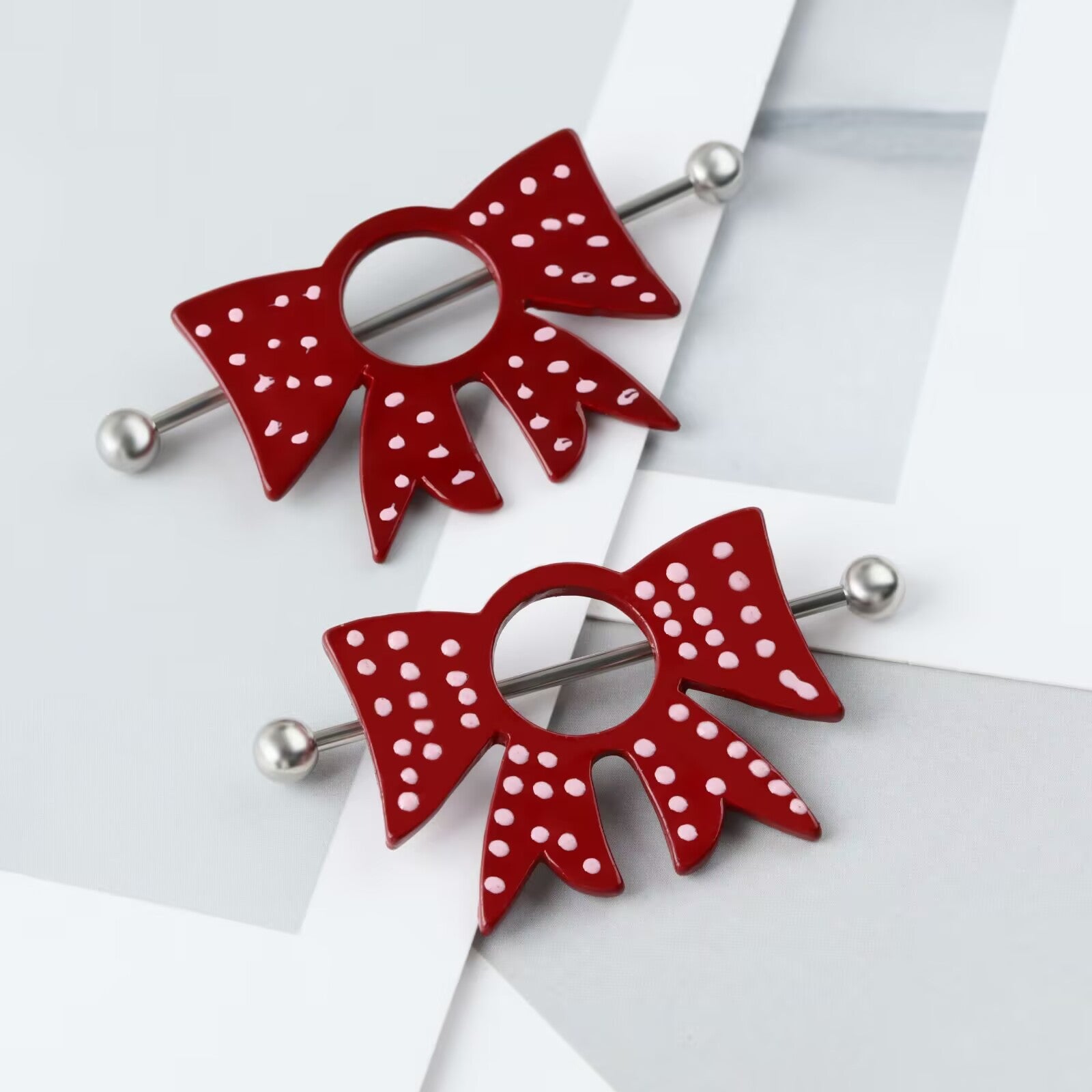Two red bowknot nipple bars with pink dots resting on a modern aesthetic grey marble and paper background.