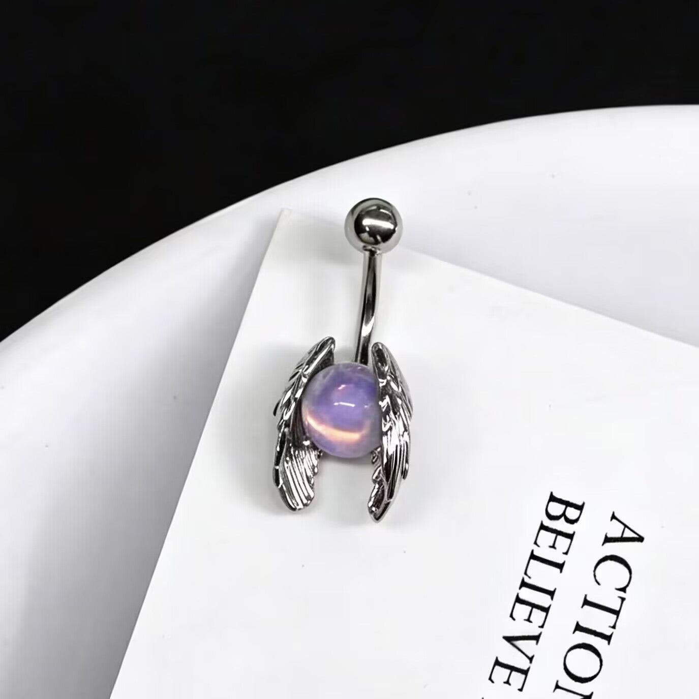 Angel wings belly button ring with a glowing purple opalite stone resting on a white card