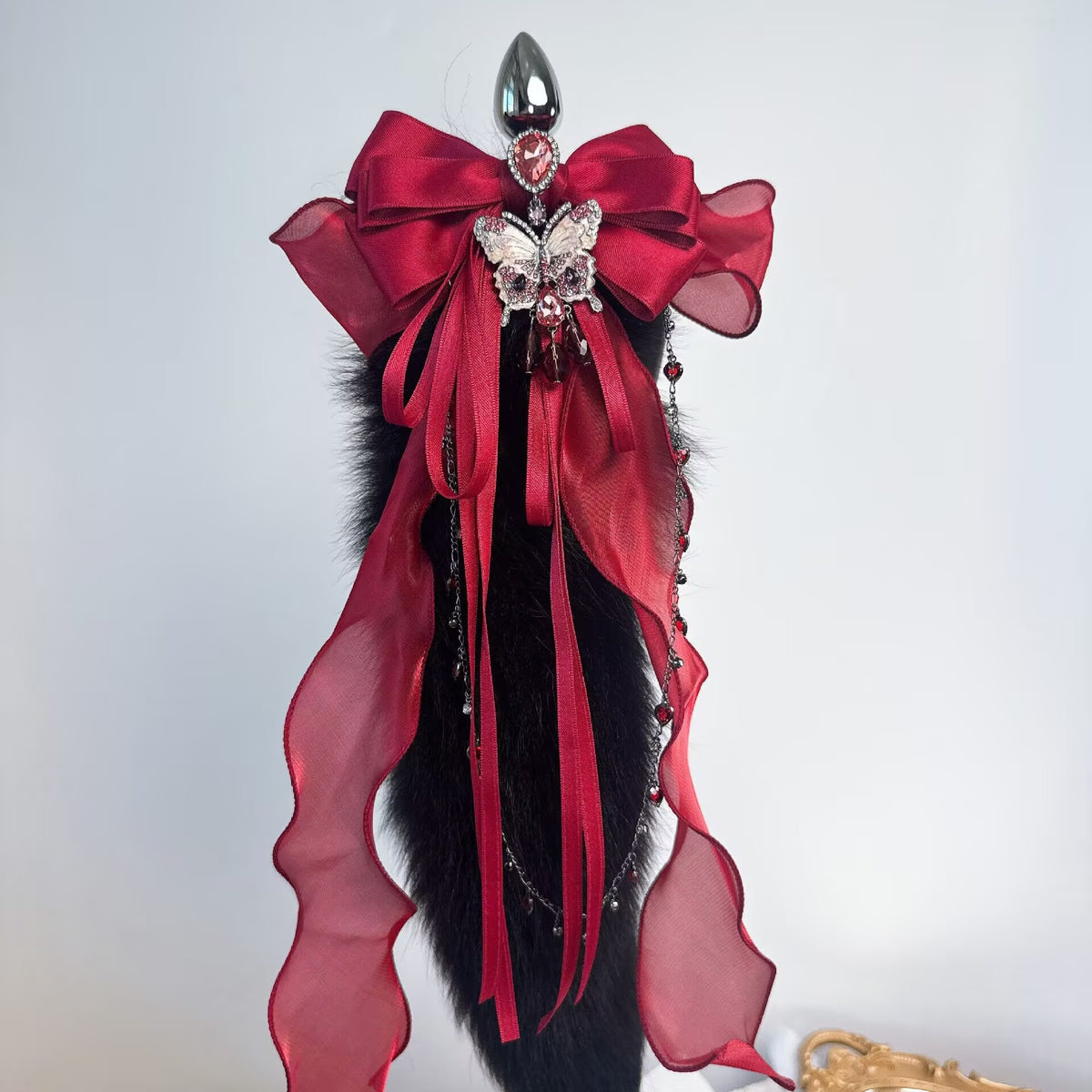 Metal anal plug with soft black faux fur tail, large red ribbon bow, and rhinestone butterfly brooch