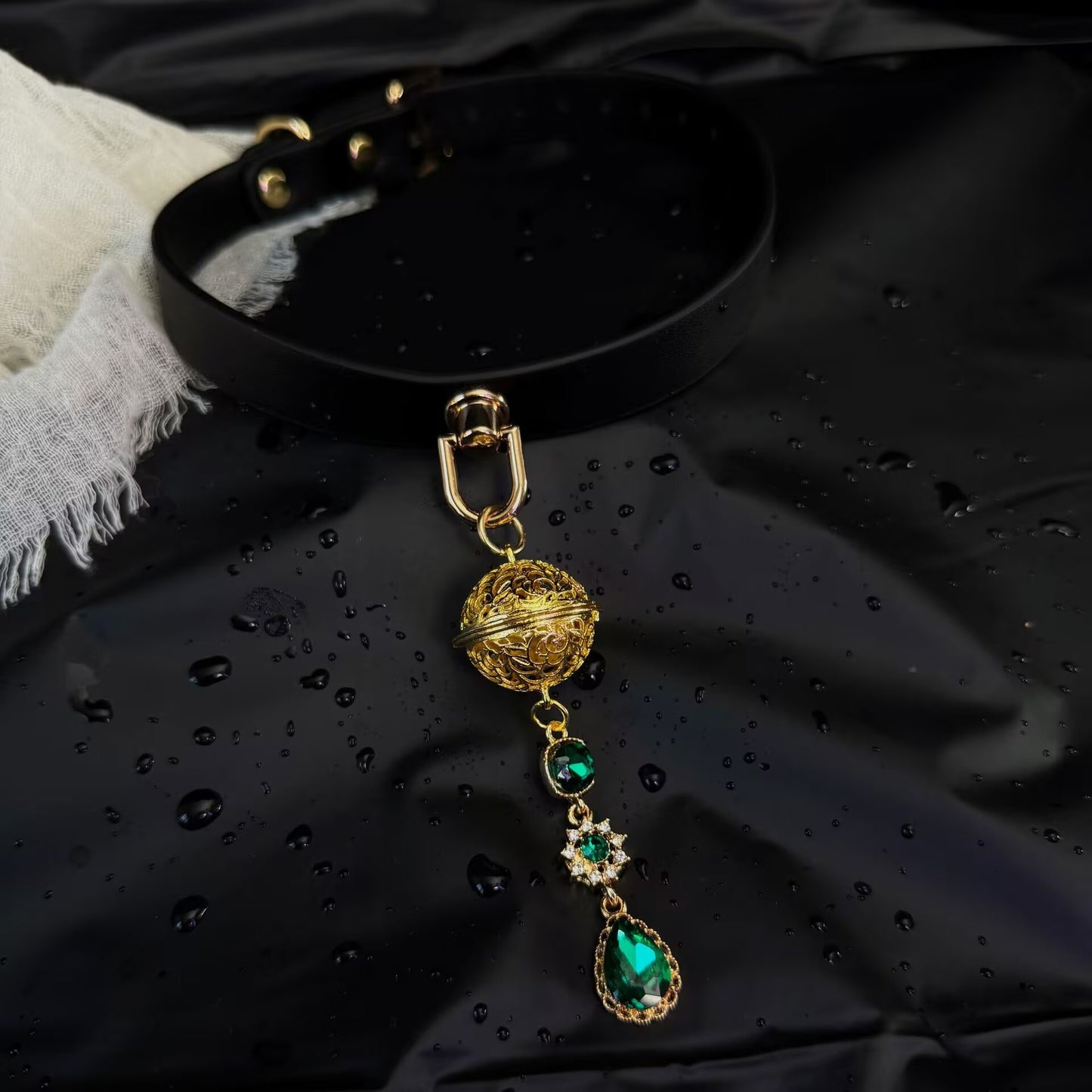 Black leather choker collar with a vintage gold filigree sphere and emerald teardrop crystal pendant on dark background.