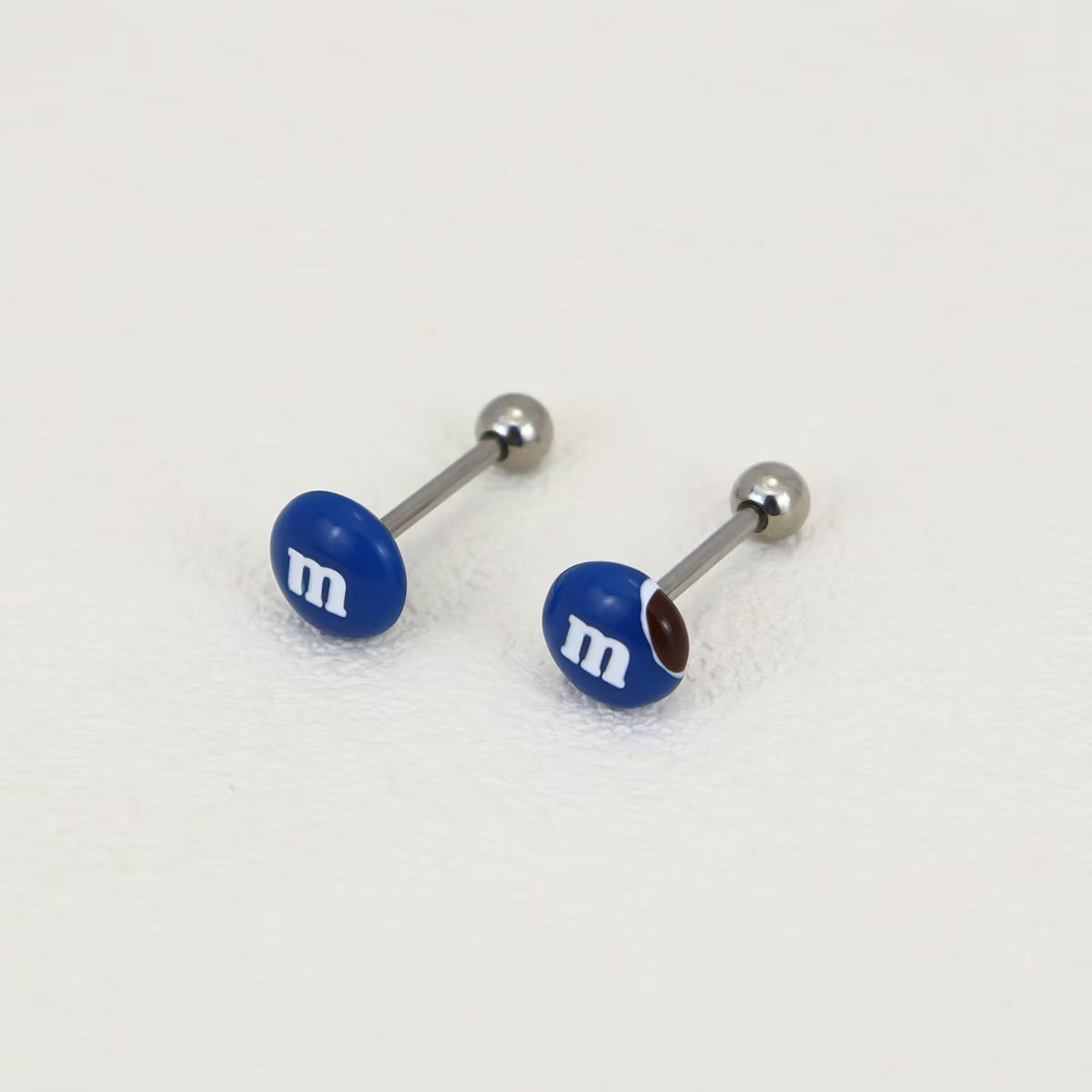 Pair of royal blue M&M candy tongue barbells with m-logo details on a light textured background.