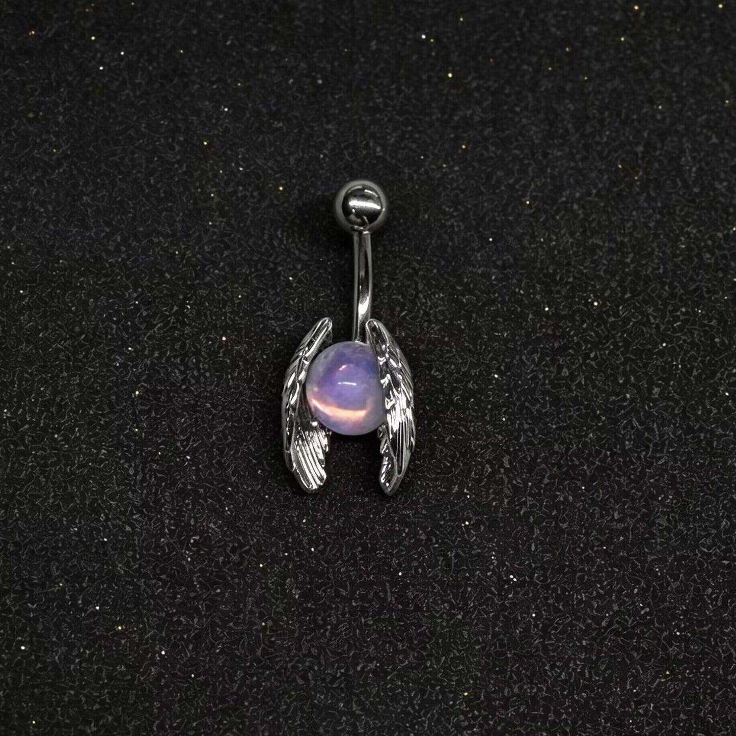 Celestial angel wing navel ring with iridescent purple stone displayed on a dark textured surface