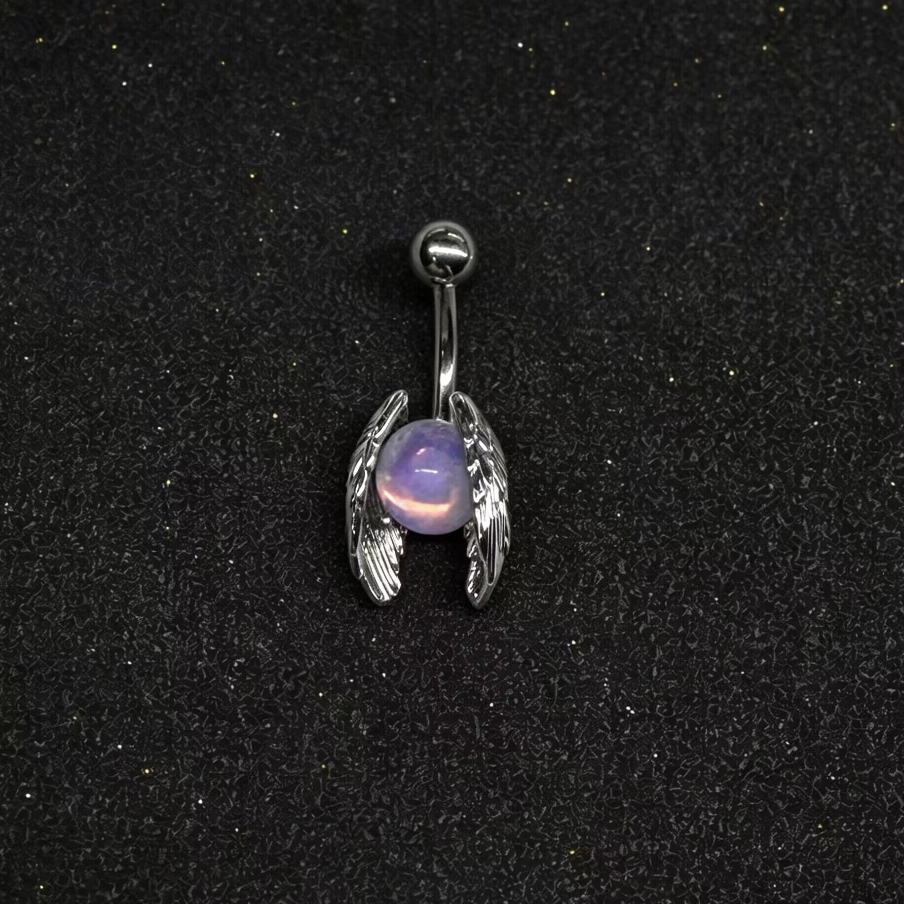 Celestial angel wing navel ring with iridescent purple stone displayed on a dark textured surface