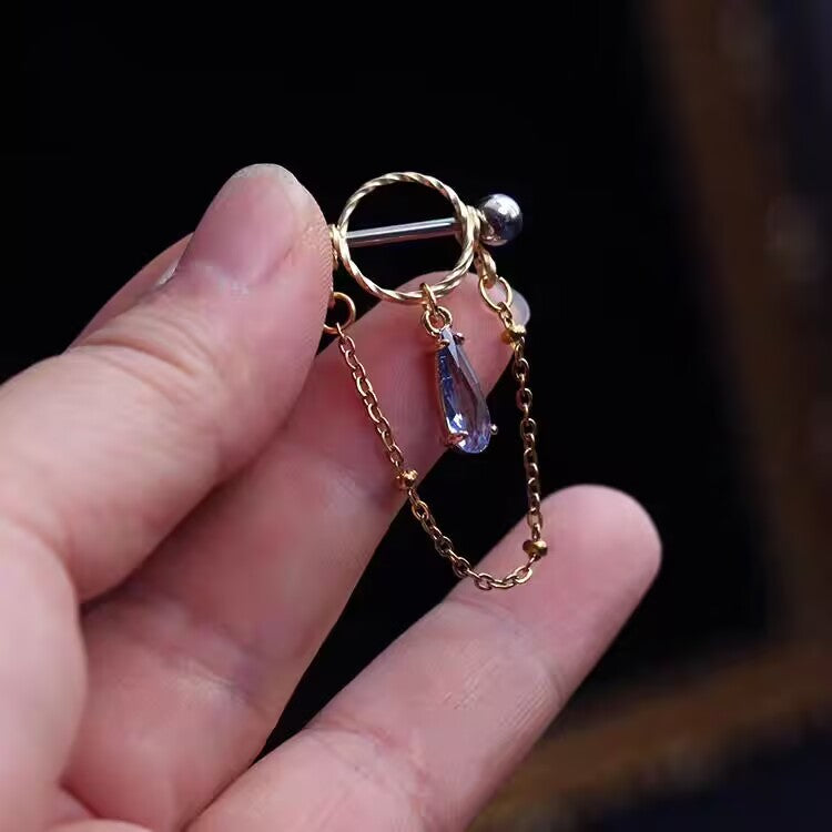 Hand holding a high quality gold nipple shield ring with a tanzanite blue waterdrop CZ pendant and chain.