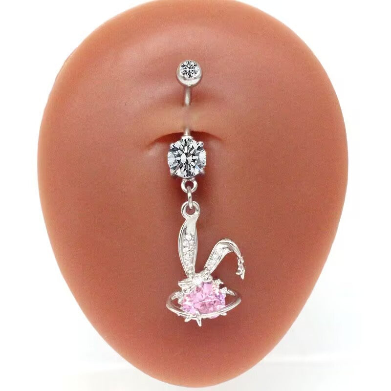 A realistic silicone navel model wearing a shiny coquette aesthetic bunny ear belly button ring with a dangling pink heart saturn pendant.