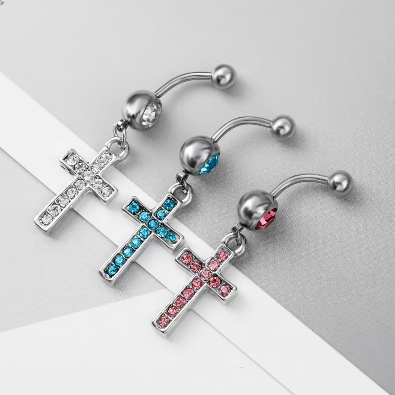 Three cross dangle belly piercing rings displayed nicely on a minimal modern background.
