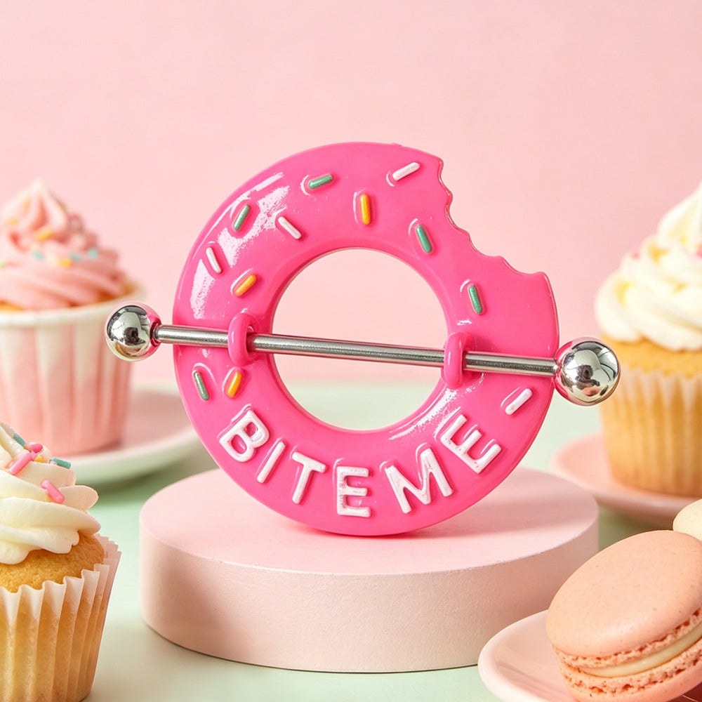 A lifestyle display of the cute pink donut nipple ring on a pedestal surrounded by cupcakes and macarons for a coquette aesthetic look.