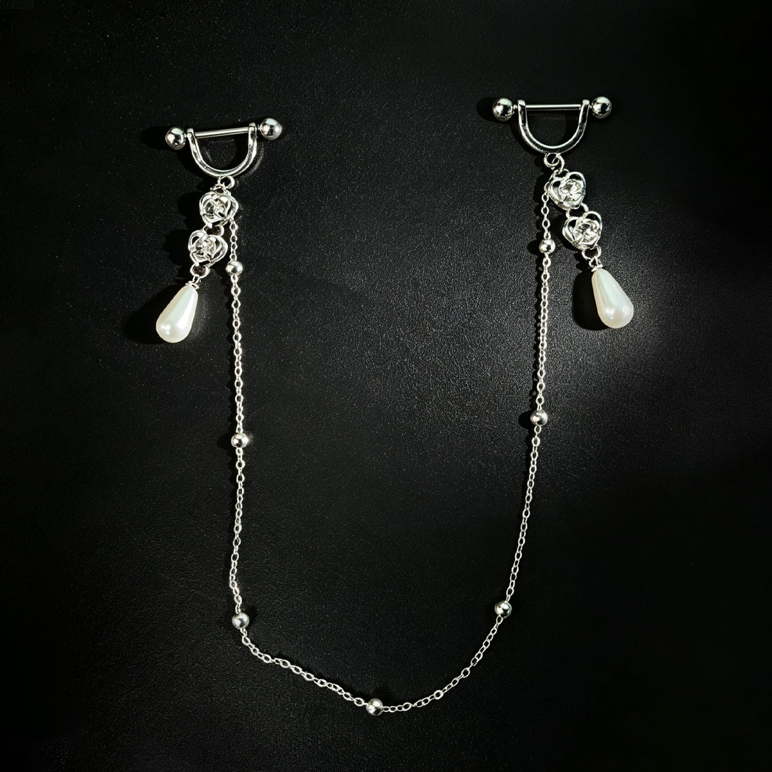Front view of a one-piece integrated body jewelry set featuring double barbell rings connected by a long drape chain with heart shapes and teardrop pendants on a dark background.