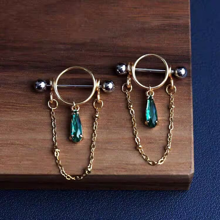 Pair of gold nipple shield rings featuring emerald green waterdrop gemstones and hanging side chains.