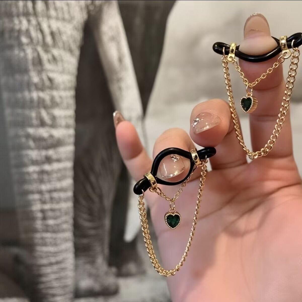 Hand showing non-pierced body jewelry featuring emerald green rhinestone heart pendants and gold tassels.