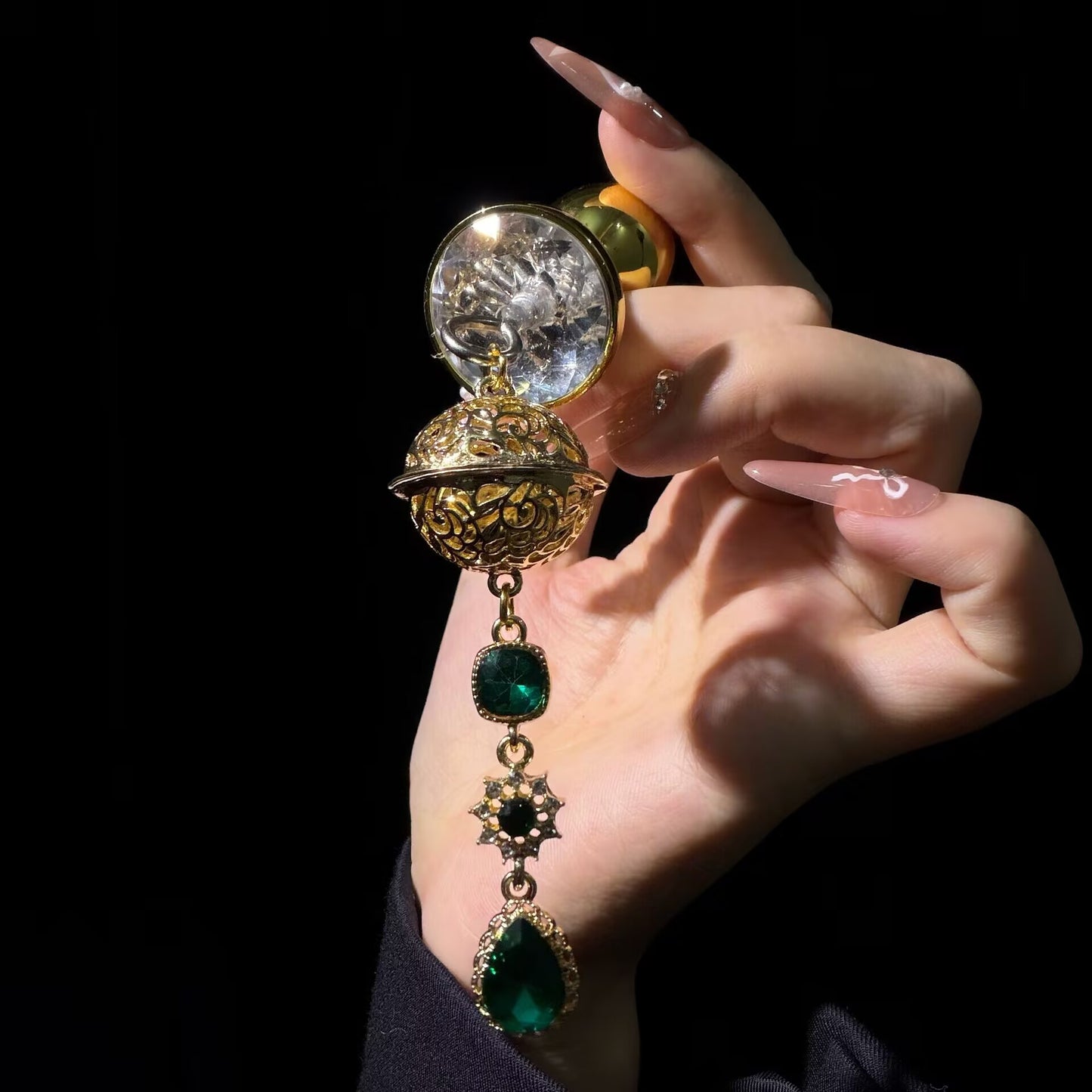 Hand holding luxury gold-toned non-pierced nipple clamp featuring a clear crystal suction cup, intricate filigree orb, and green emerald gemstone dangle charms.