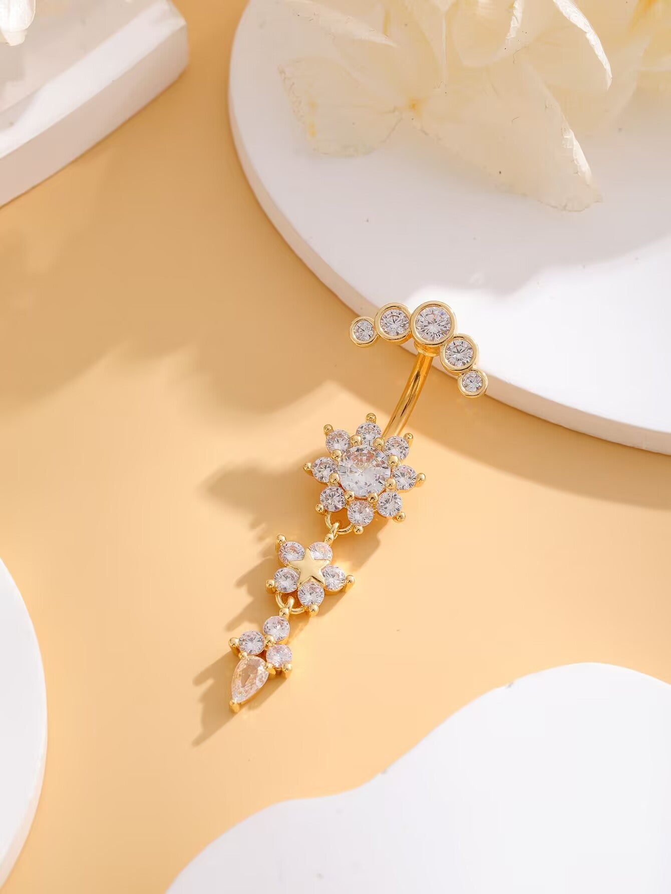 Flat lay of gold floral CZ dangle belly ring with clear cubic zirconia accents on soft background.
