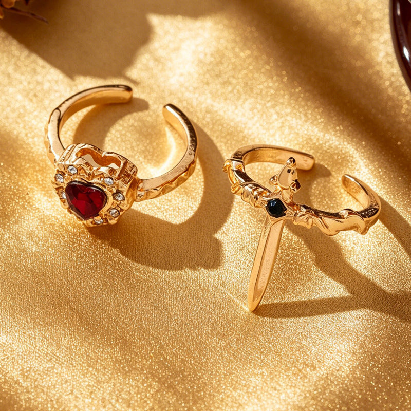 One gold-tone red heart open ring and one black-accent sword open ring on gold fabric