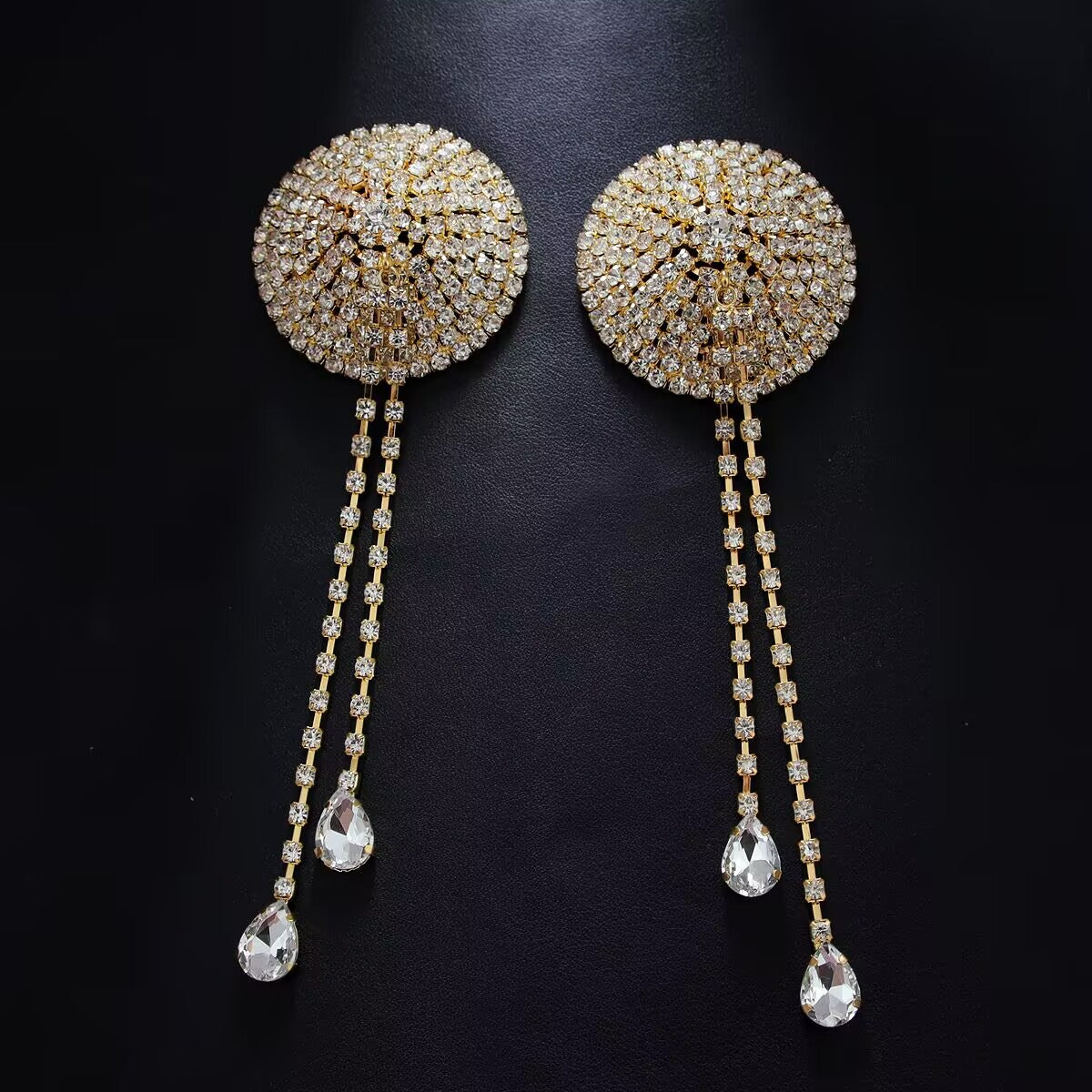 Pair of gold full-coverage rhinestone nipple covers with dangling crystal tassels on dark textured background.