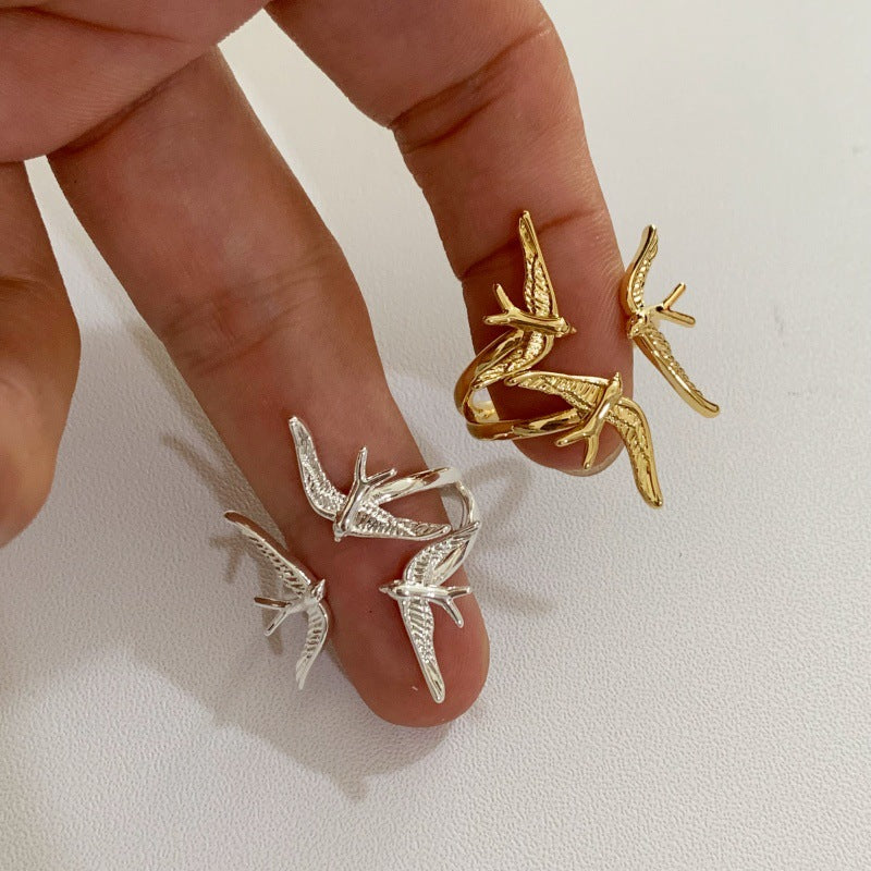 Gold and silver-tone swallow rings held on fingertips, showing their bands and raised bird motifs.