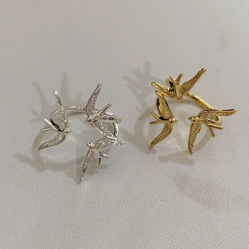 Two three-swallow rings, one silver-tone and one gold, on a light textured surface.