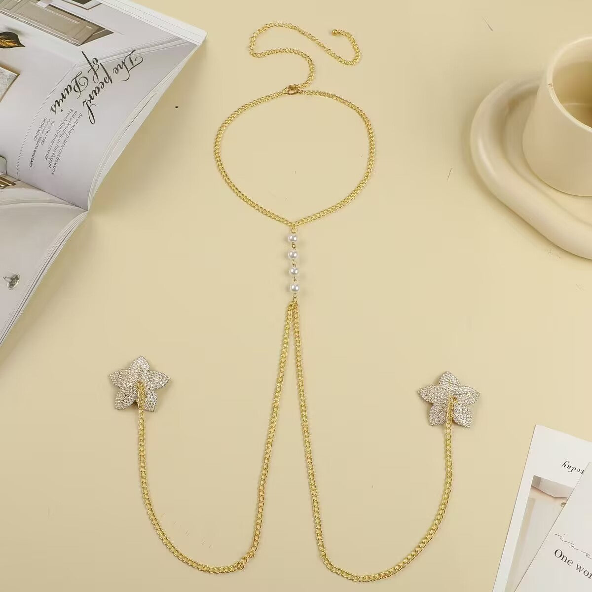 Flatlay of a luxury gold non pierced nipple shield chest chain with rhinestone star motifs and elegant central pearl beads on a warm beige background.