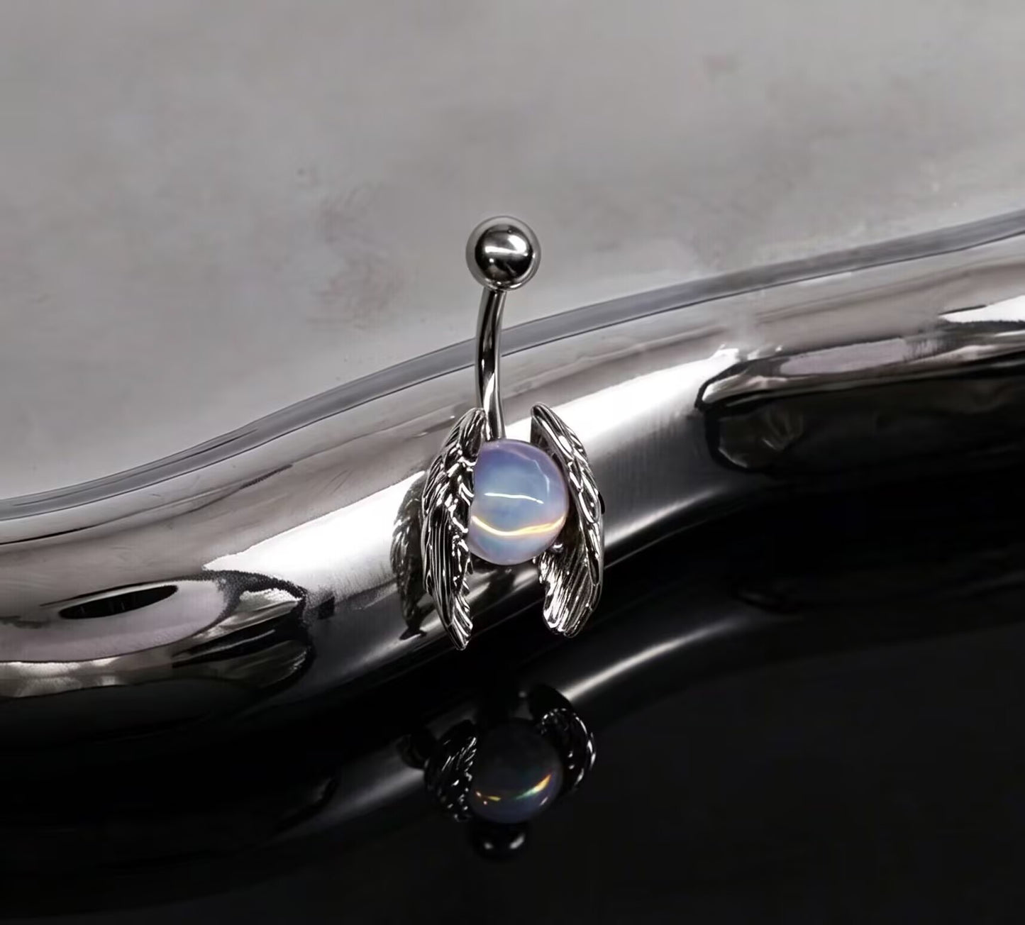 Gothic angel wings belly ring with iridescent orb reflecting elegantly on a metallic surface