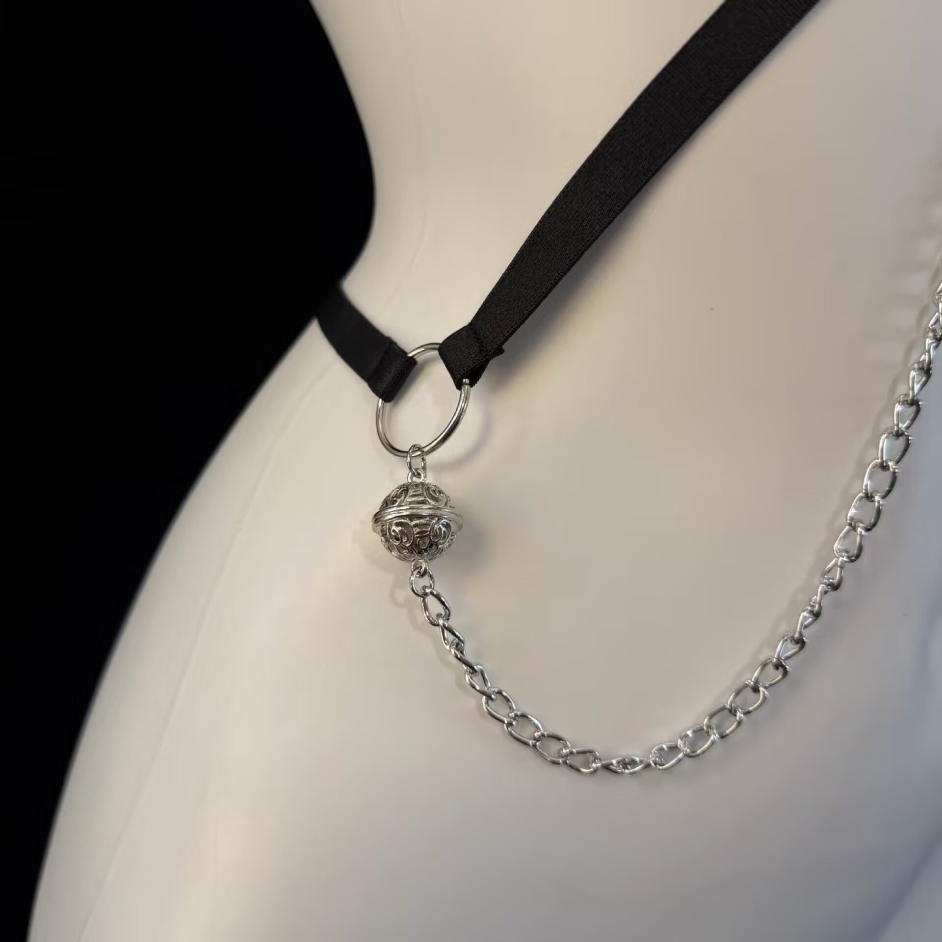 Close-up detail of silver chain connector with vintage hollow orb charm and black fabric strap on body harness.