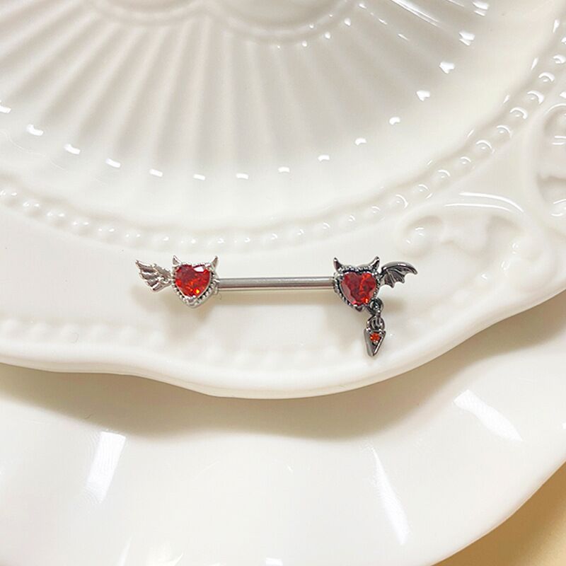 Aesthetic display of a devil winged nipple ring with a ruby red heart on a decorative white ceramic plate.