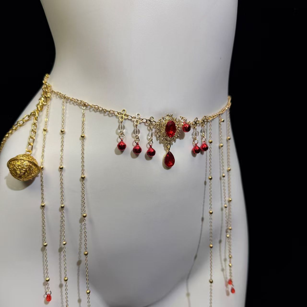 Close-up view of the gothic royal gold belly chain on a white mannequin, highlighting the intricate red ruby cameo pendant, dangling tassel chains, and a hanging filigree gold bell charm.