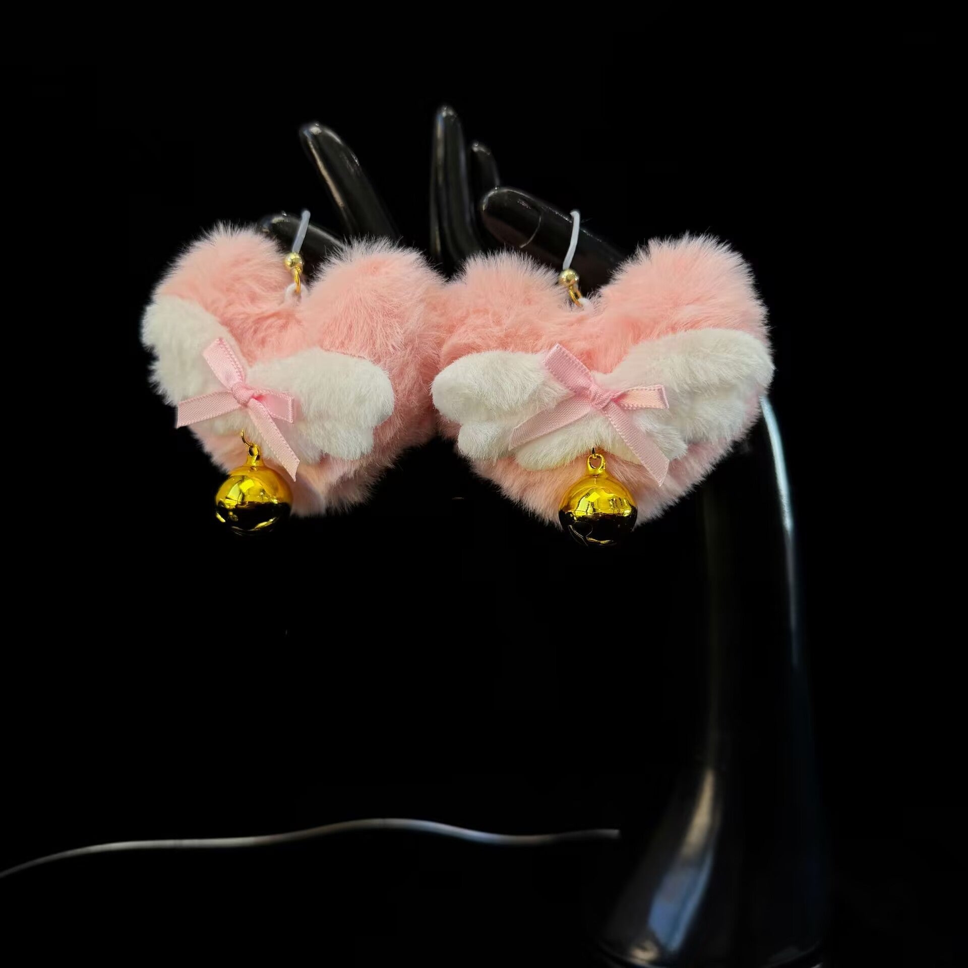 Pair of kawaii pink heart nipple clamps with white angel wings and jingle bells displayed on a mannequin hand against a black background.