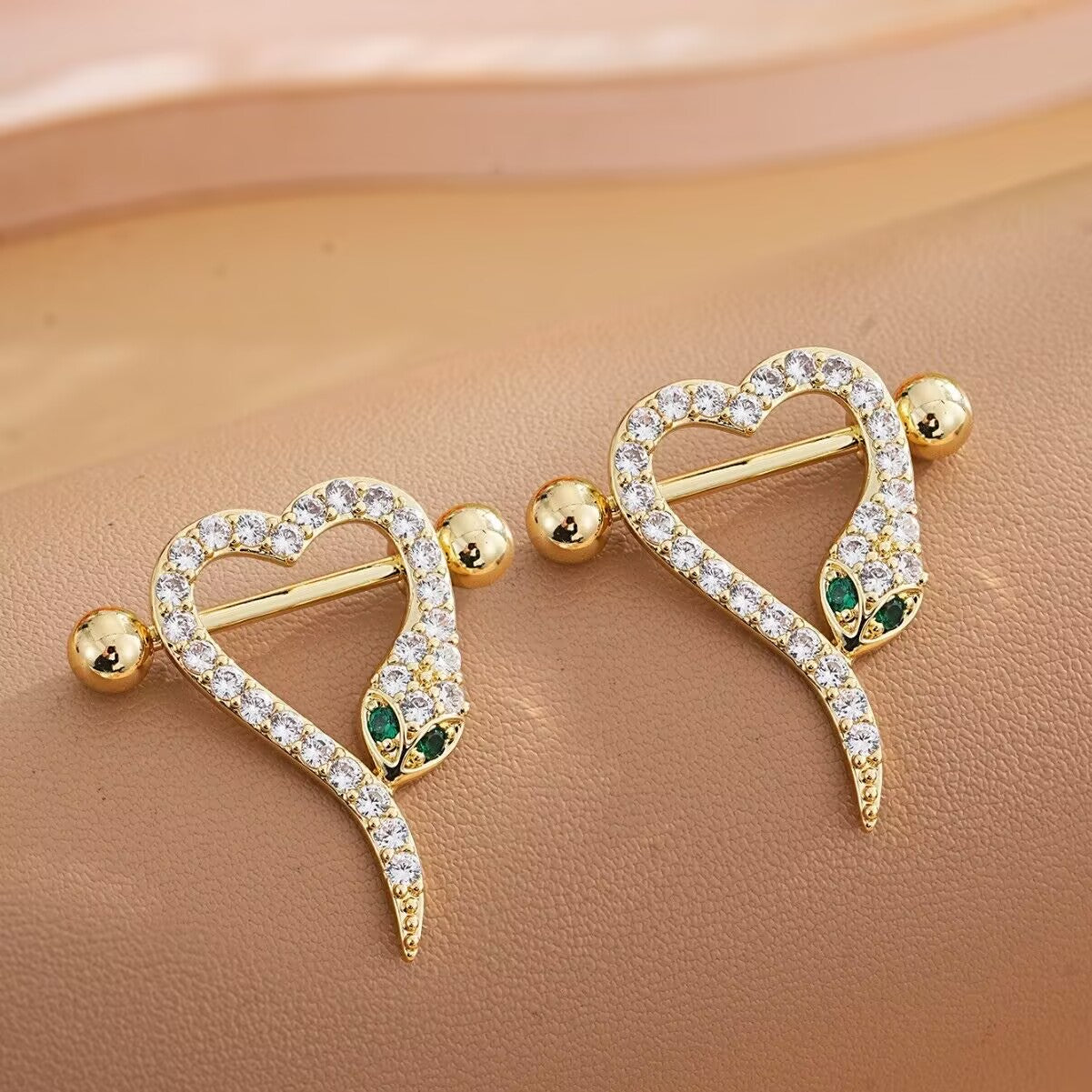 Angled lifestyle shot of gold snake heart nipple rings on a soft leather background.