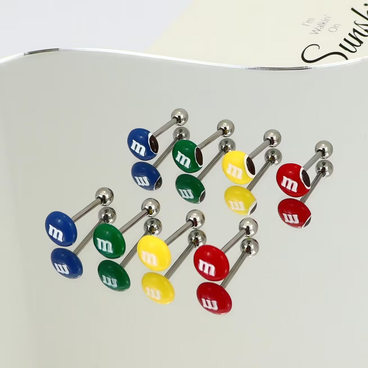 A collection of colorful M&M candy tongue barbells with solid and bitten designs reflecting on a clean surface.