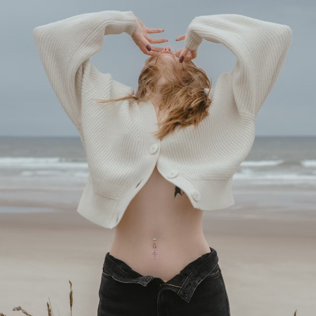 Aesthetic photoshoot of a female model wearing a pink cross dangle belly button ring at the beach.