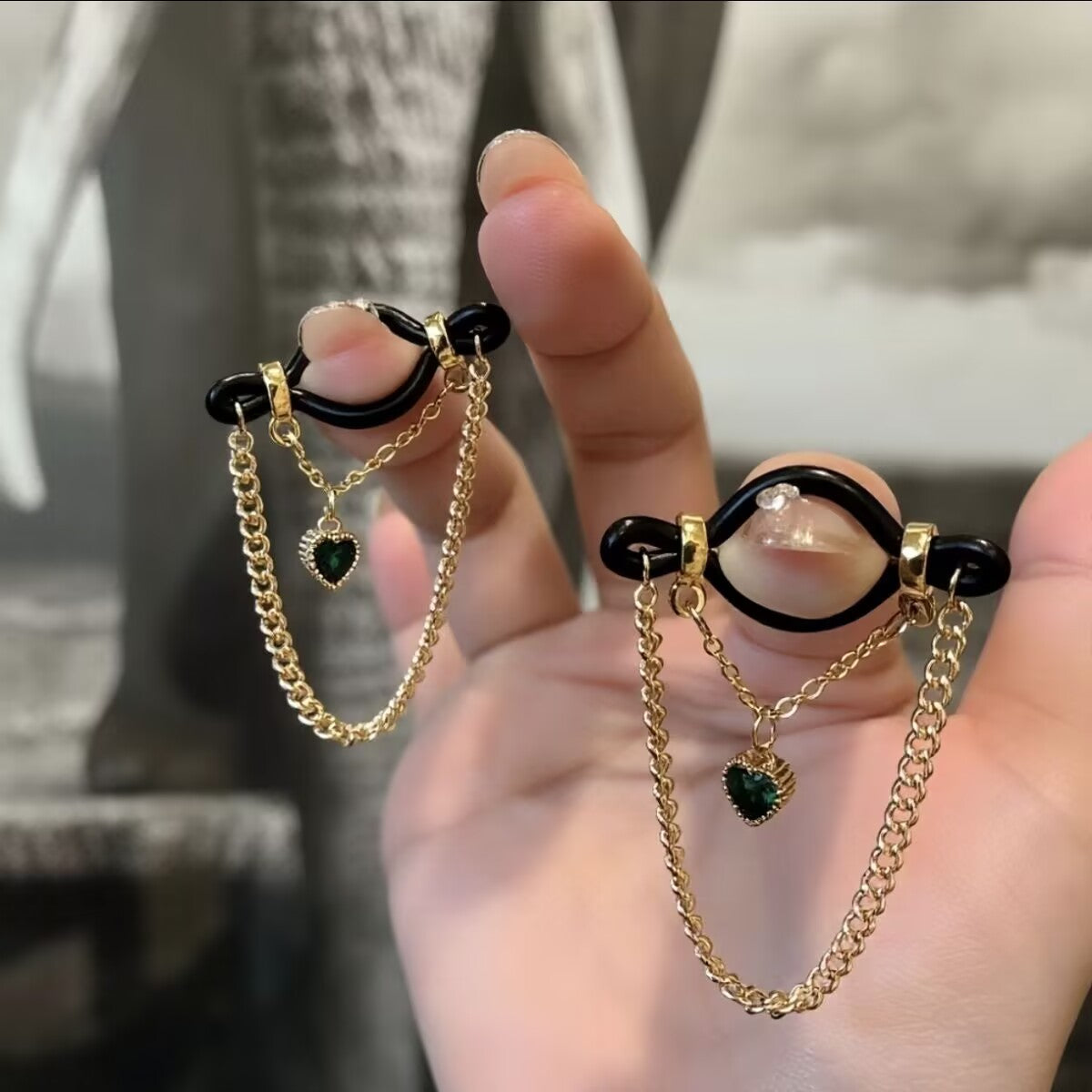 A pair of non-pierced gold nipple ring clamps with double layer chains and green crystal heart charms held by a hand.