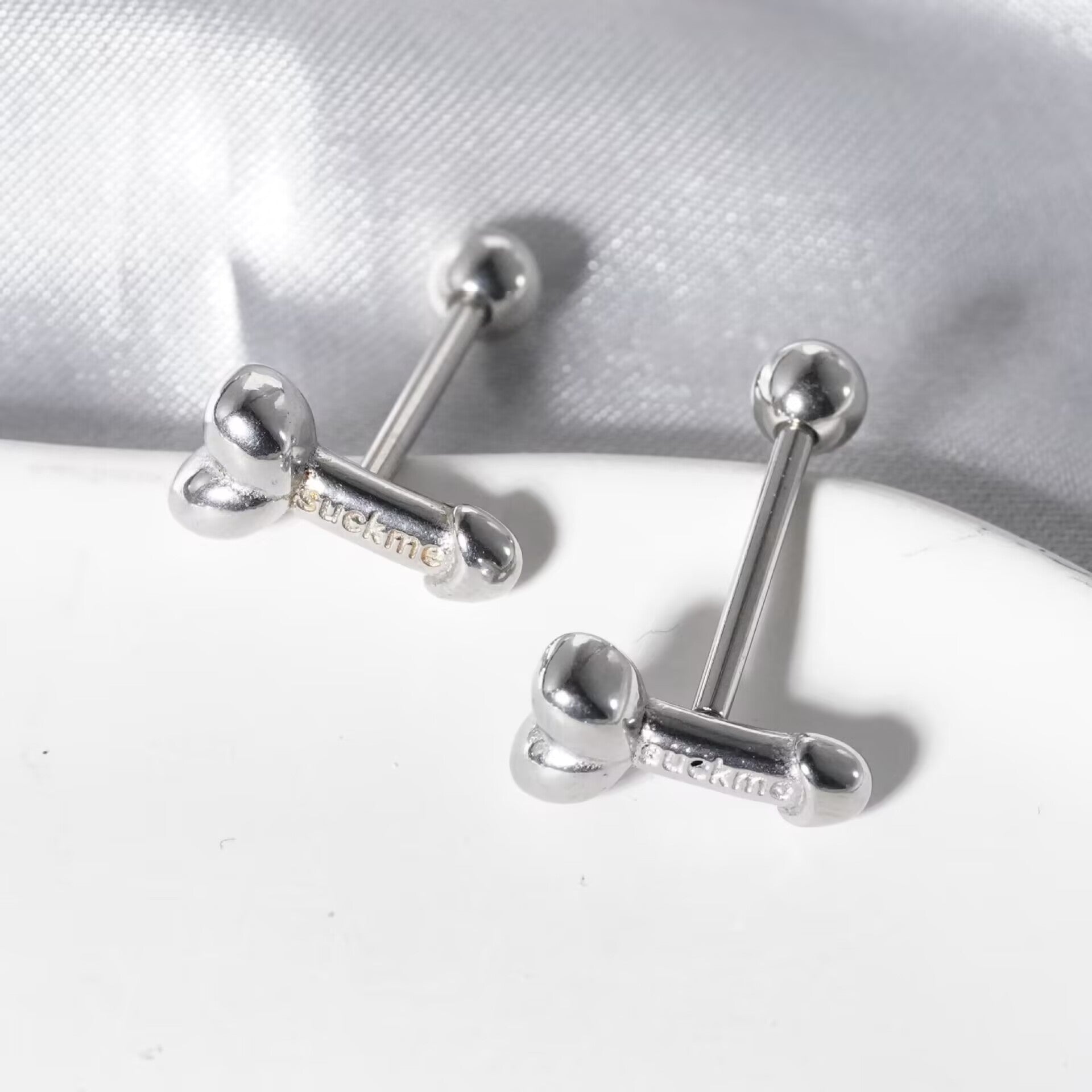 Close-up of silver penis-shaped tongue rings with engraved suck me lettering resting on a display tray.