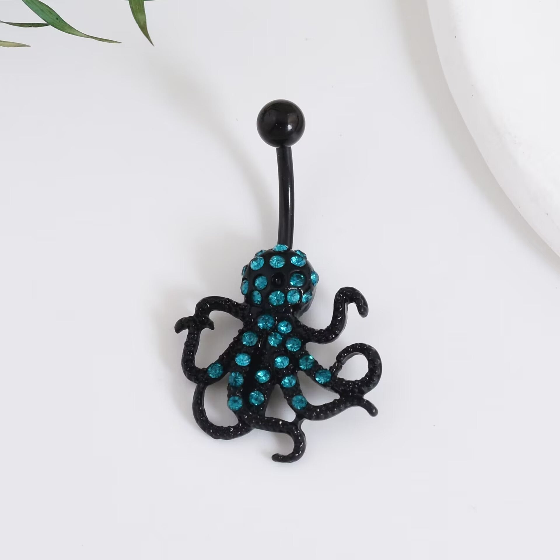 An ocean punk style black kraken navel ring with shimmering teal blue crystals on a reflective white surface.