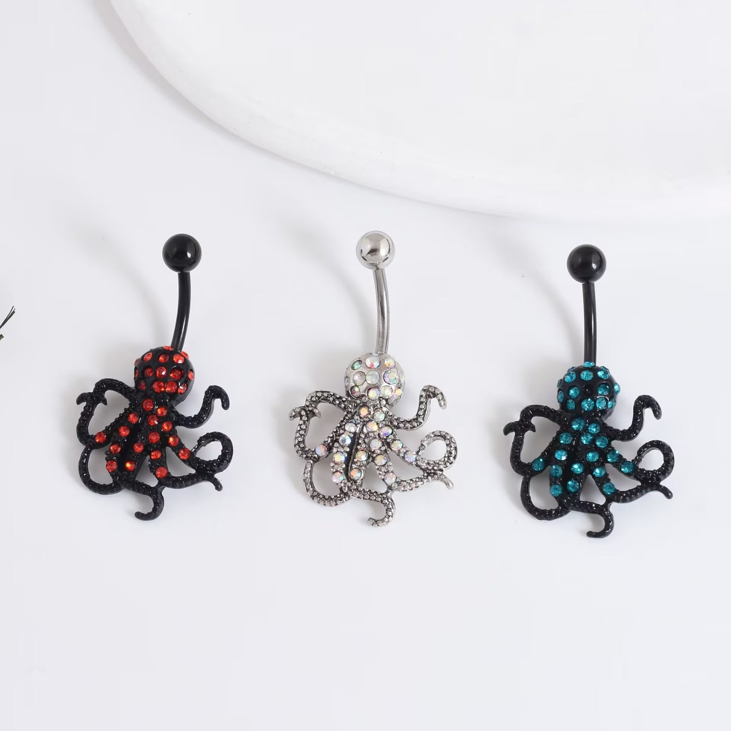 Three variations of the alternative octopus navel piercing jewelry in red rhinestones, iridescent AB crystal, and teal blue stones.