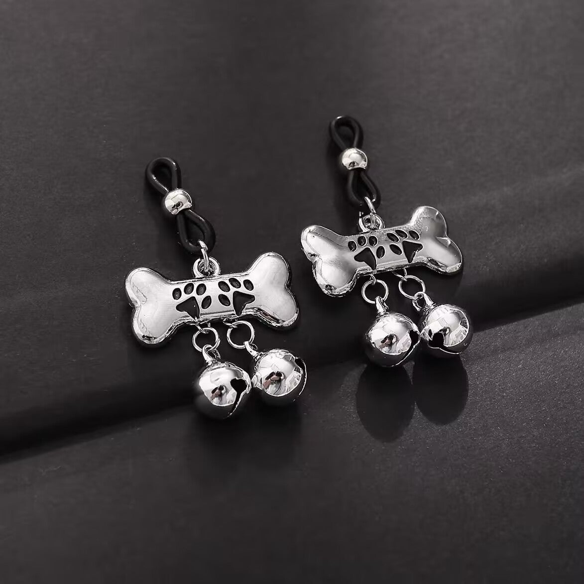 A pair of silver bone-shaped nipple loops with dangling bells on a dark textured surface.