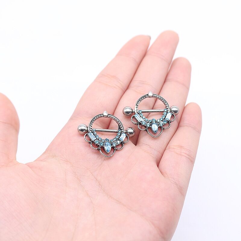 A pair of ornate blue gemstone crown nipple shields held in a hand for size comparison.