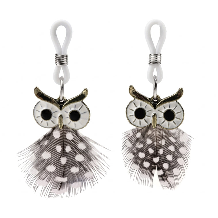 A pair of non-pierced metal nipple clamps with intricate owl designs and black and white spotted feather dangles, suspended from white silicone loops. This jewelry is designed for non-invasive body adornment.