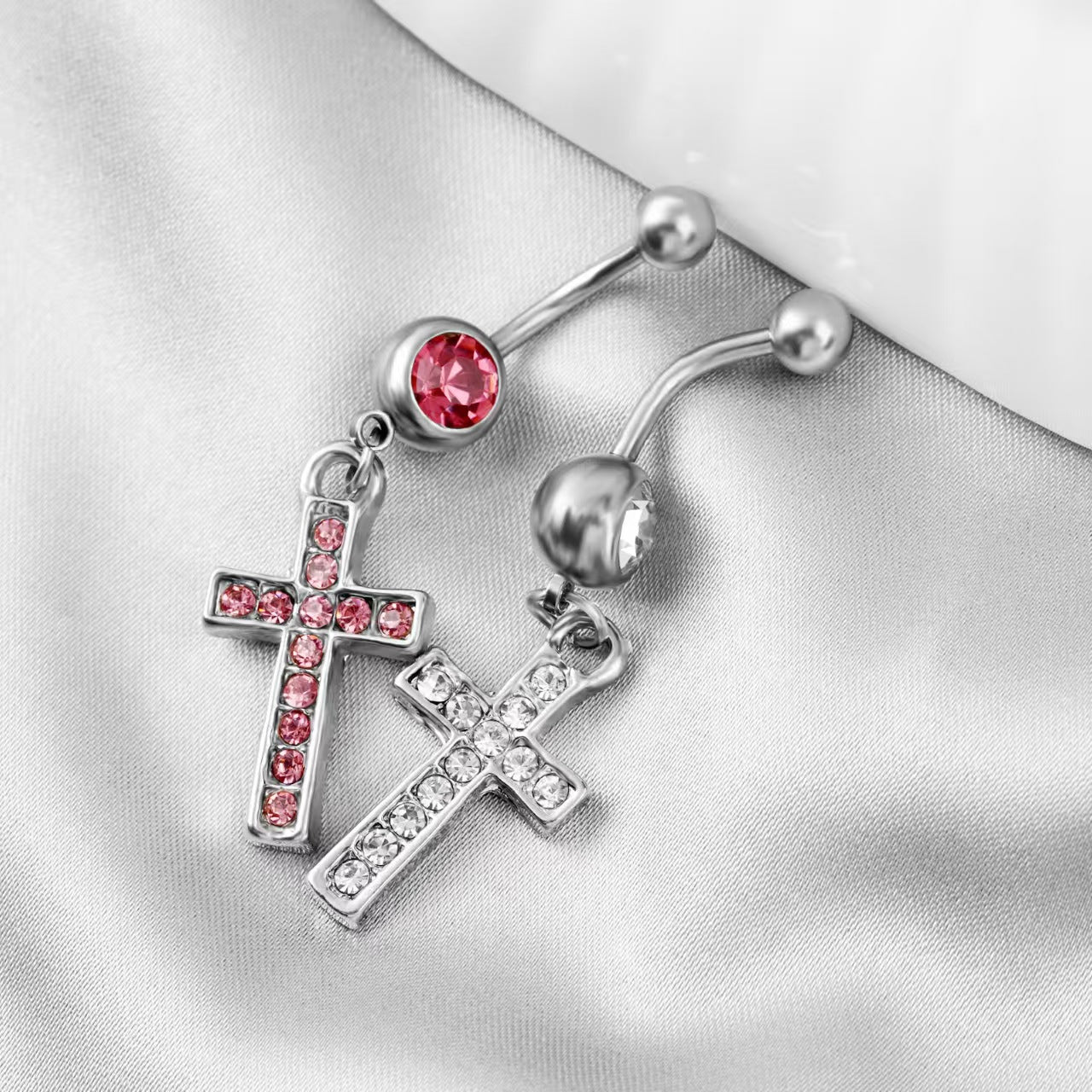 Silver-colored dangling cross navel jewelry in pink and clear colorways displayed on a white fabric.
