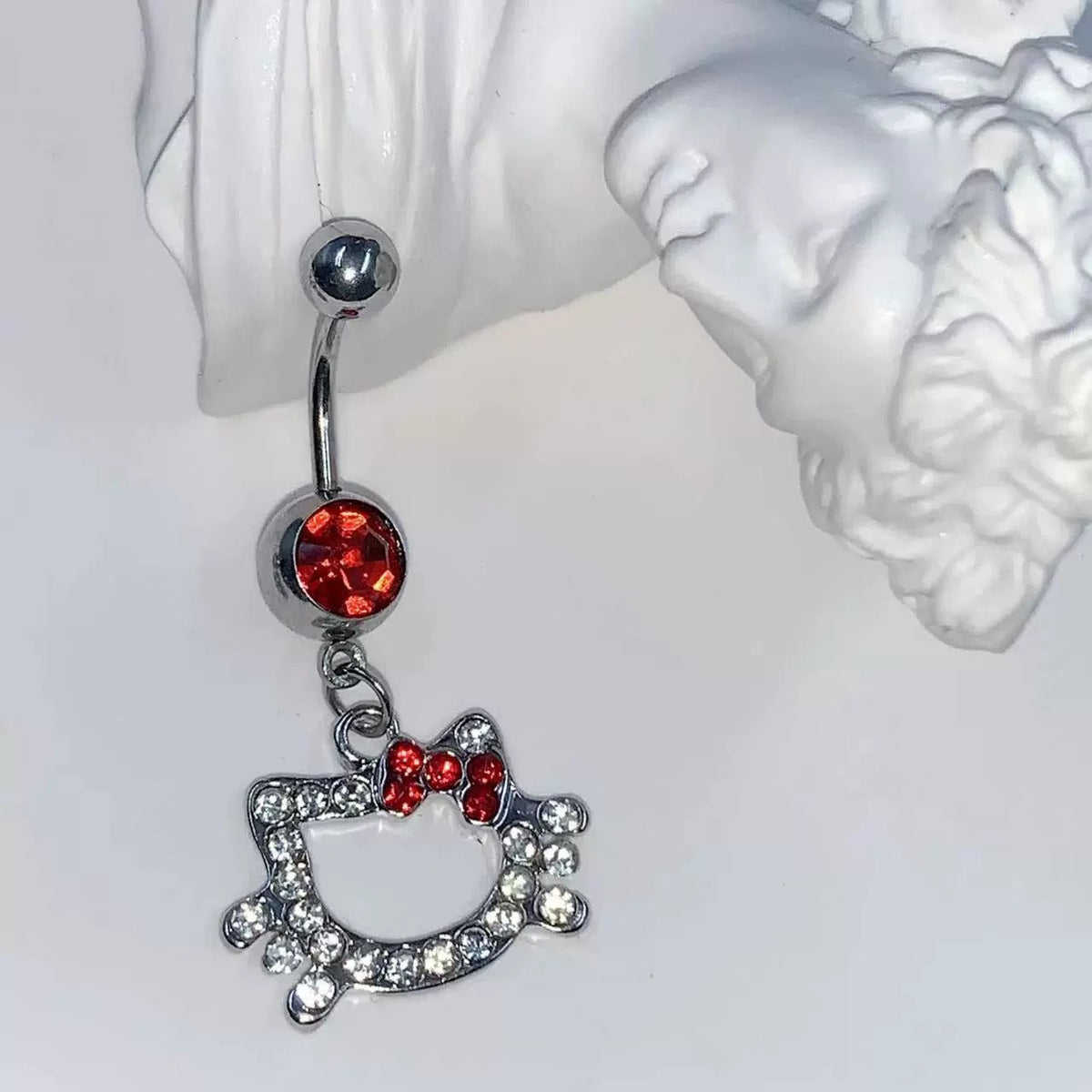 Single silver-tone red cubic zirconia cat-and-bow belly button ring beside a white sculptural ornament